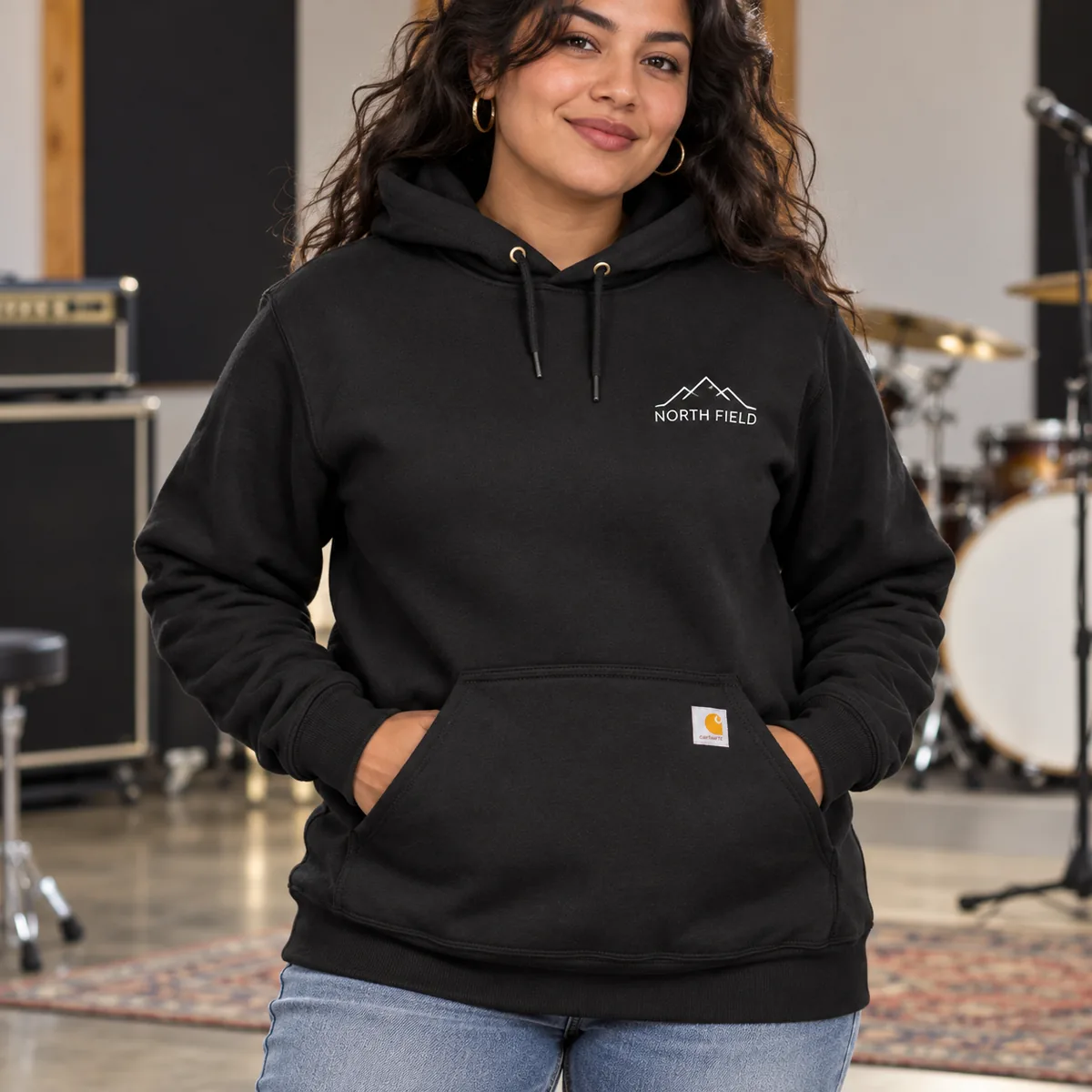 Woman wearing a black Carhartt Loose-Fit Midweight Hoodie with fictional North Field sample branding in a rehearsal studio.