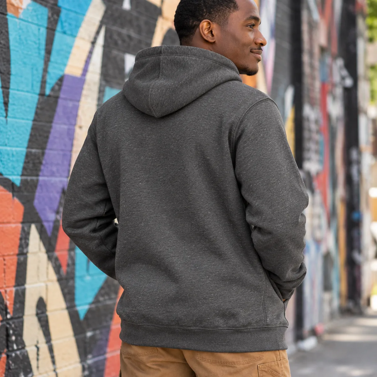 Man wearing a carbon heather Carhartt Loose-Fit Midweight Hoodie, shown from behind in a mural alley.