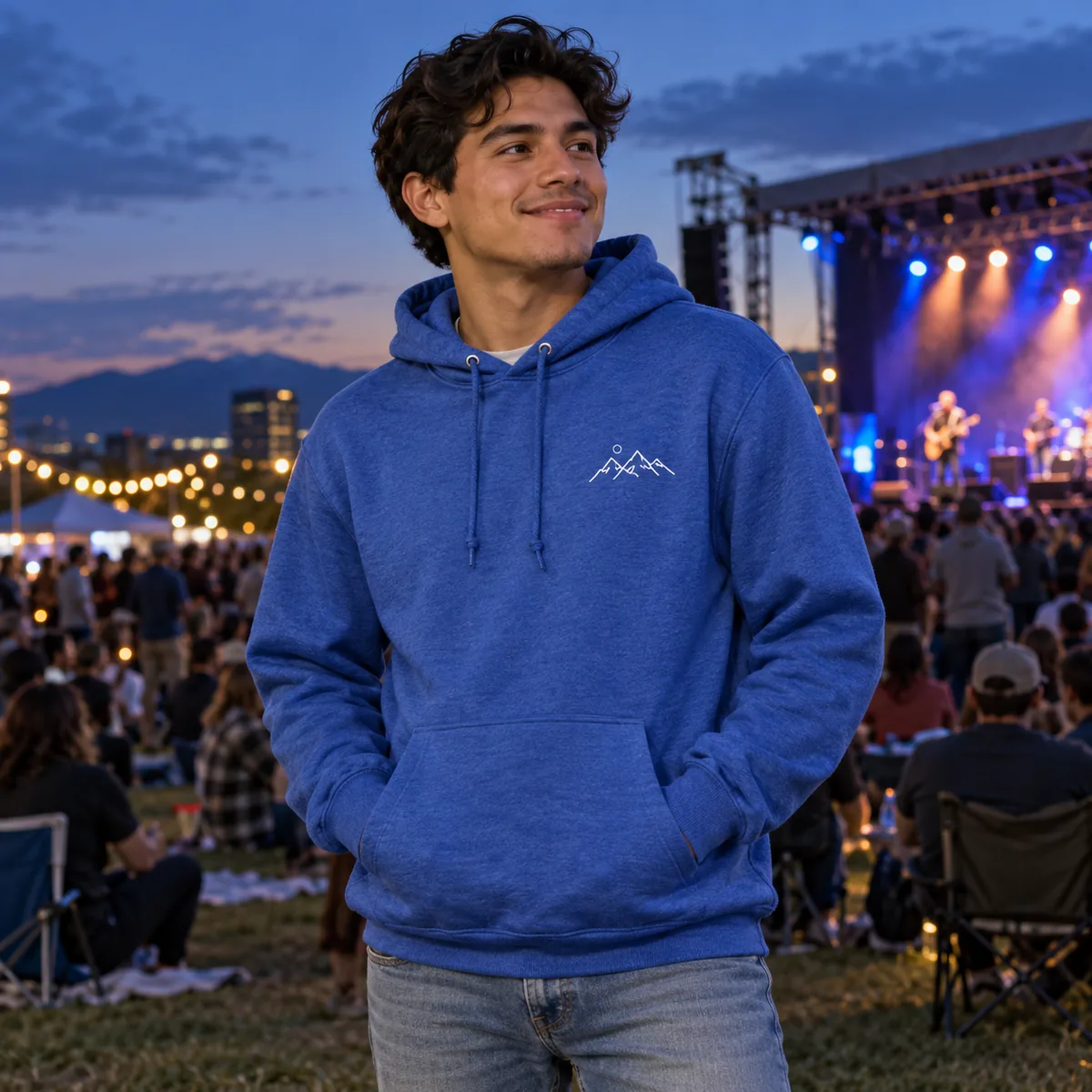 Latino man wearing a royal blue heather hoodie with fictional white mountain line art at an outdoor community concert.