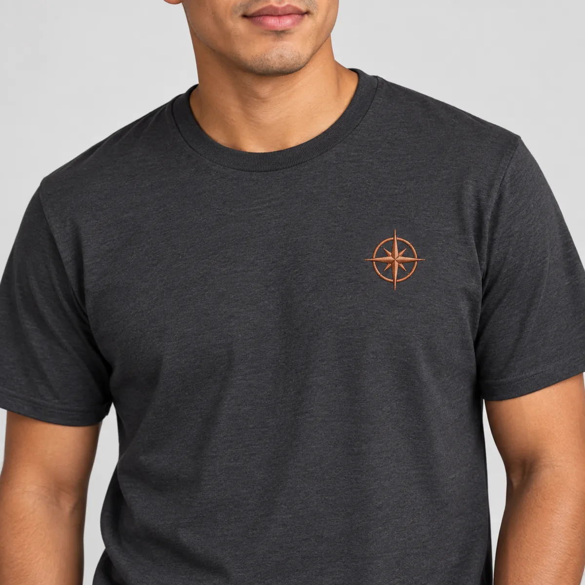 Close-up of a fictional copper compass emblem on a charcoal District Very Important Tee worn by a model.