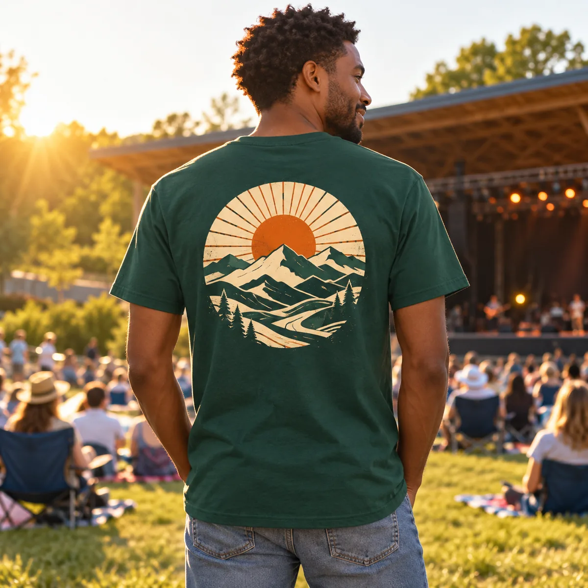 Back view of a model wearing an evergreen District Very Important Tee with fictional mountain-sun back artwork at a music pavilion.