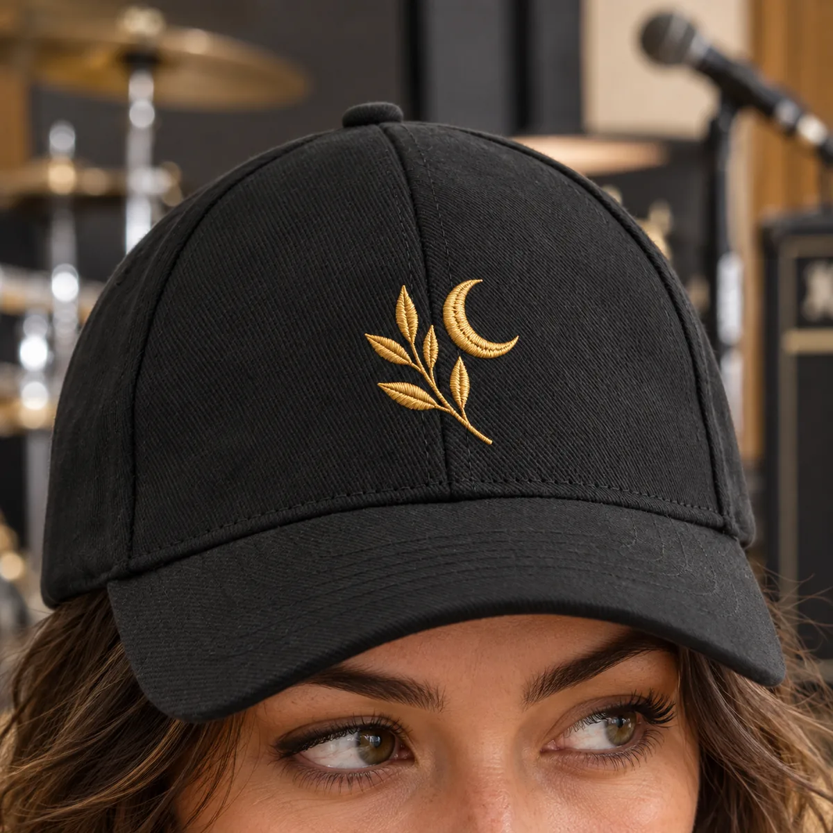 Close-up of a woman wearing a black cap with a small fictional gold embroidered leaf-and-moon emblem at a rehearsal studio.