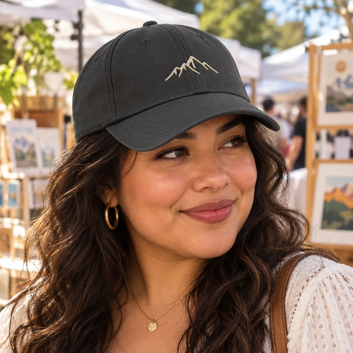 Woman wearing a dark grey cap with a fictional cream mountain embroidery at an outdoor art market.