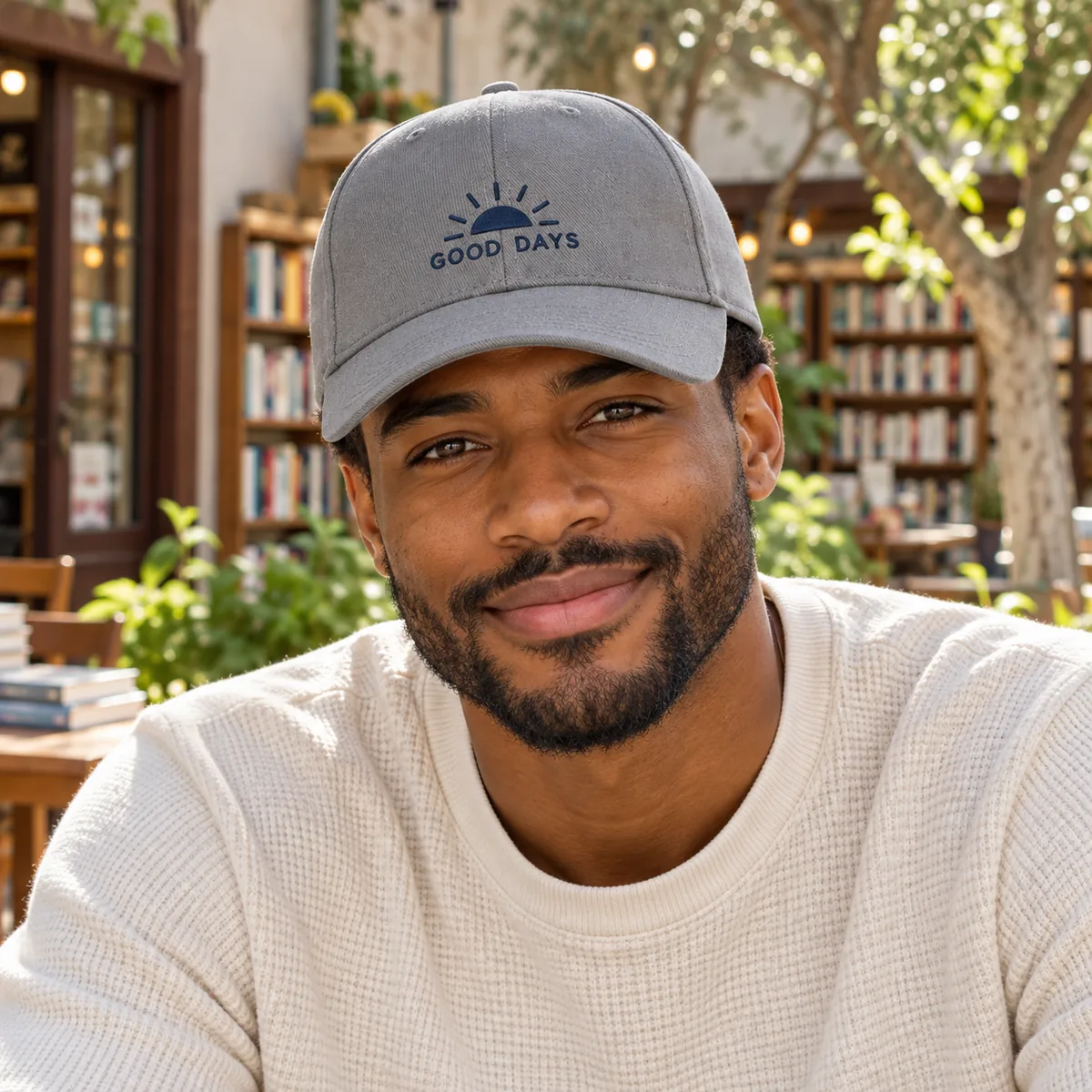 Man wearing a medium grey cap with fictional Good Days embroidered branding in a bookstore courtyard.