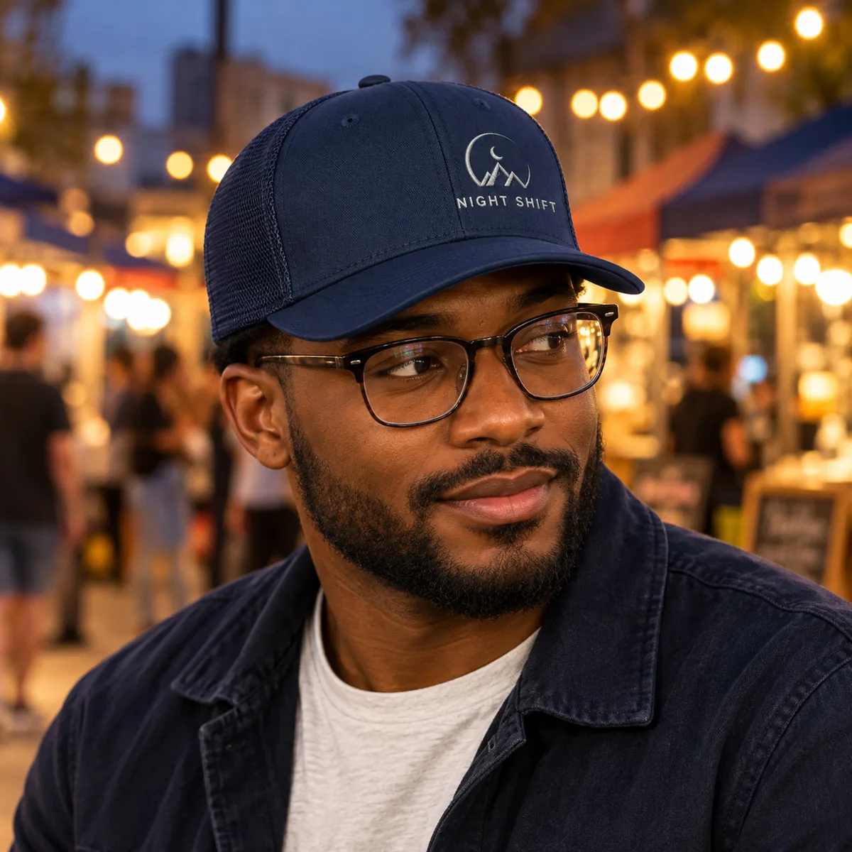 Man wearing a dark navy trucker cap with fictional Night Shift sample branding at an evening market.