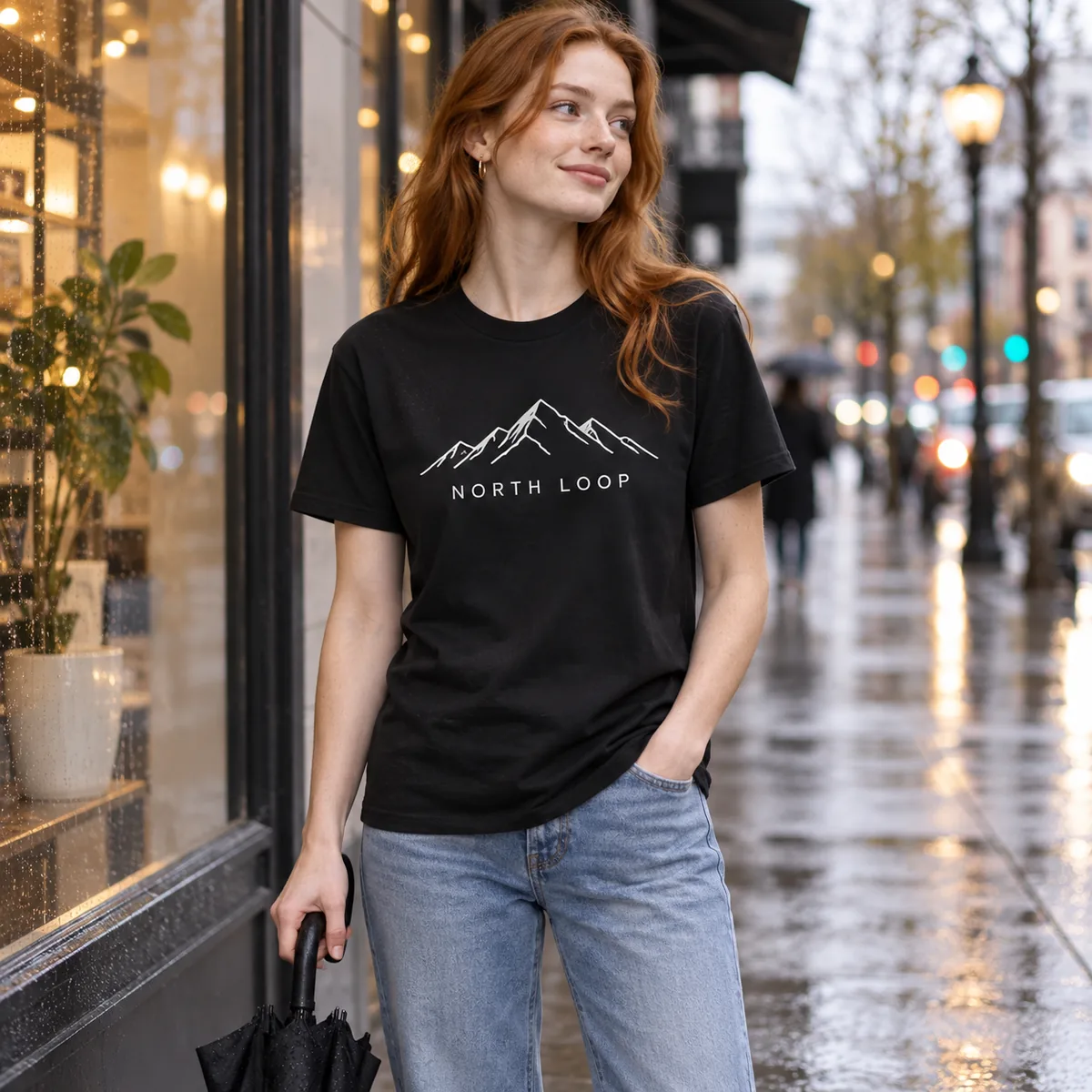 Model wearing a black Gildan Budget unisex T-shirt with fictional North Loop sample branding on a rainy city street.