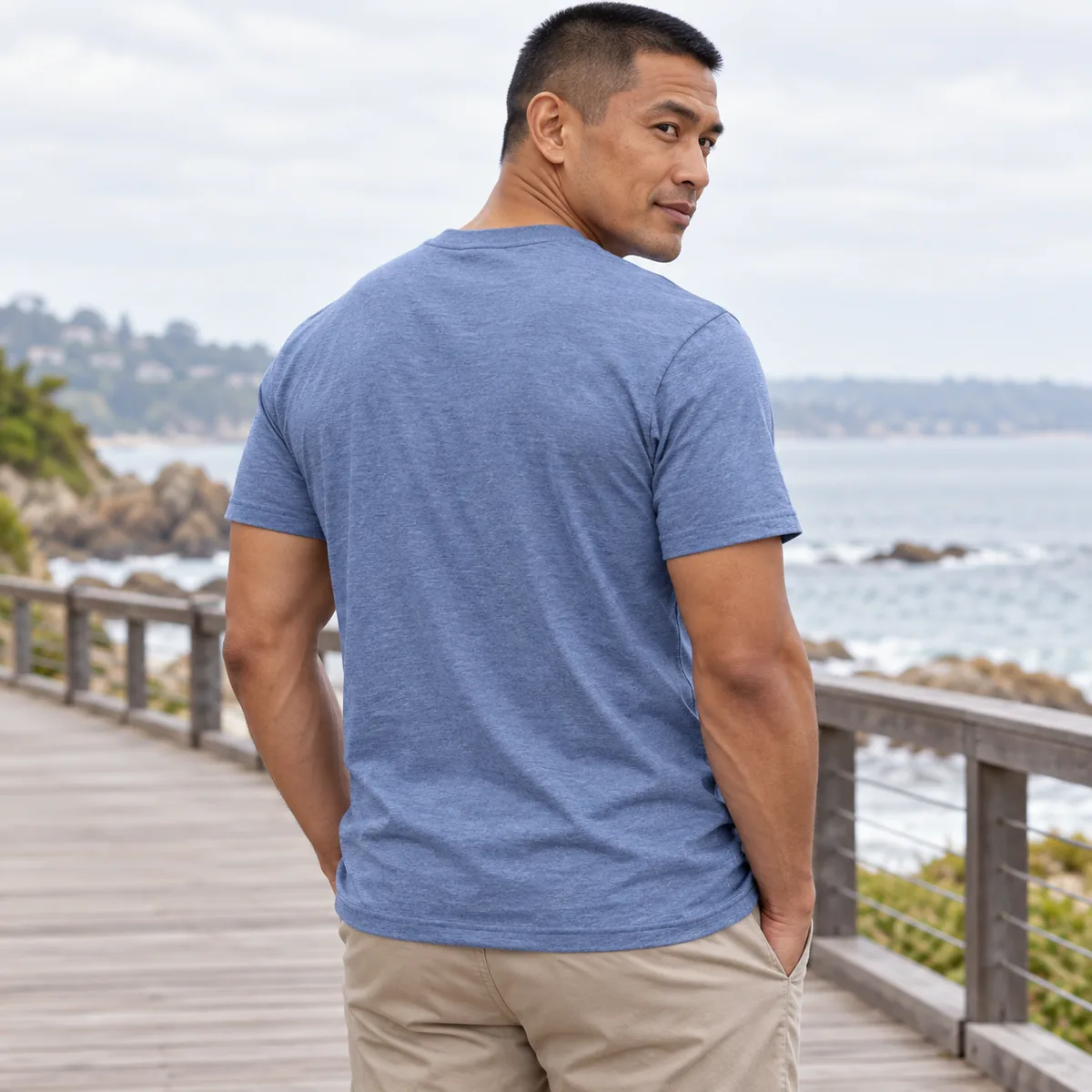 Back view of a model wearing the heather blue Gildan Budget unisex T-shirt on a coastal boardwalk.