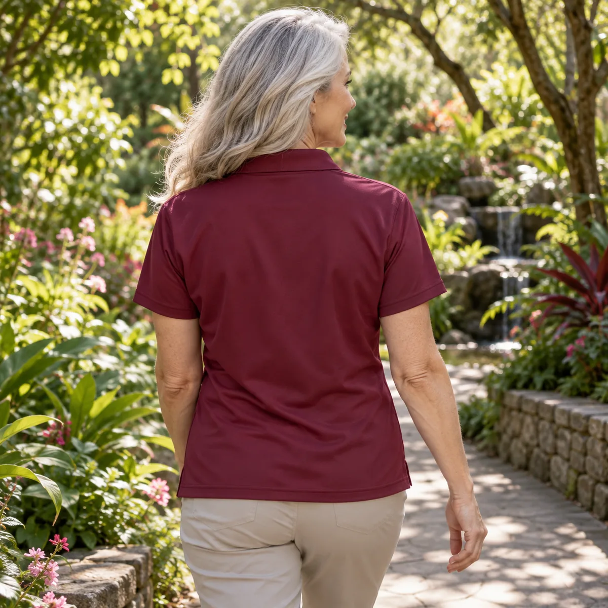 Silver-haired woman wearing a maroon Gildan DryBlend sport shirt, shown from behind in a botanical garden.
