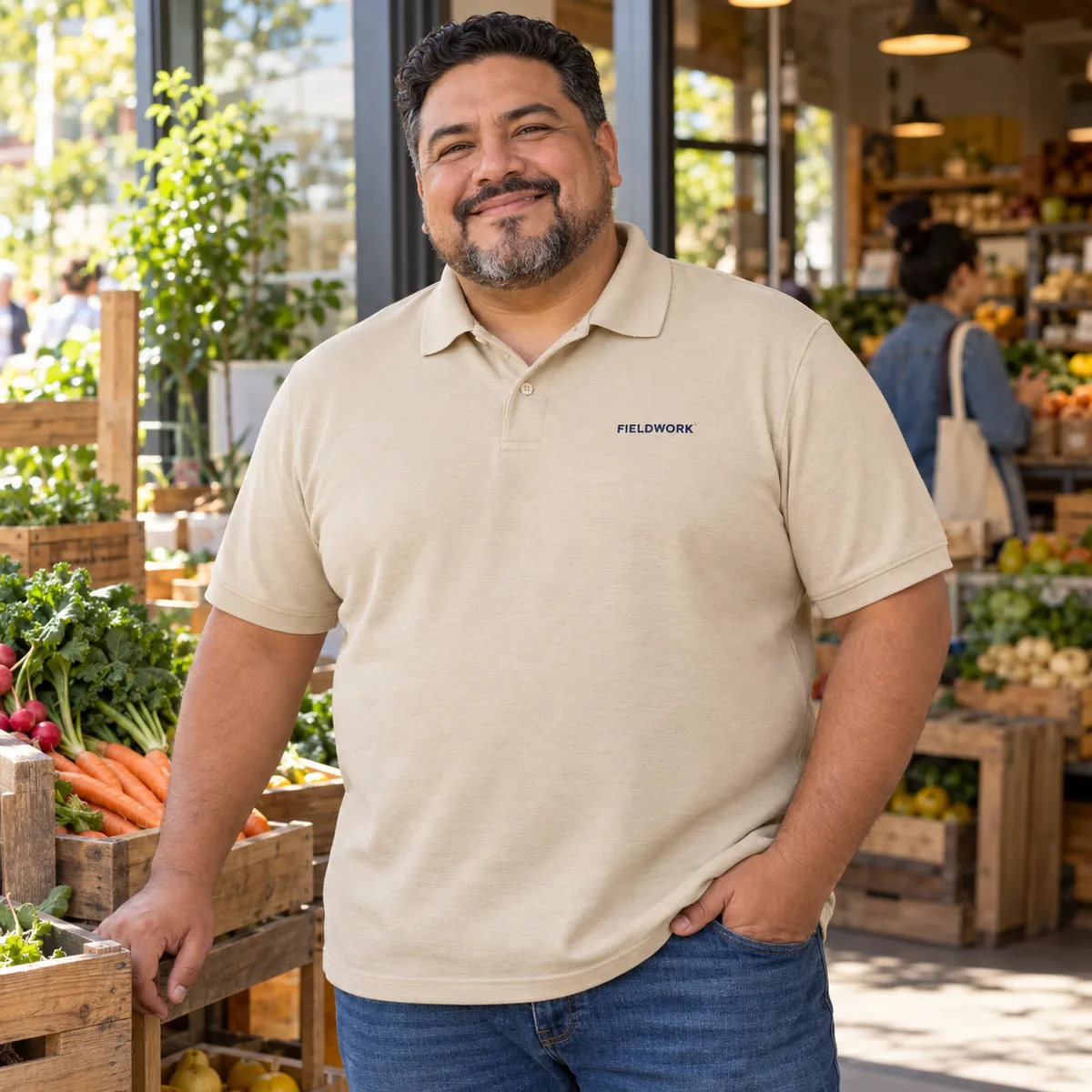 Plus-size Latino man wearing a sand Gildan DryBlend sport shirt with fictional Fieldwork chest embroidery at a community food co-op.