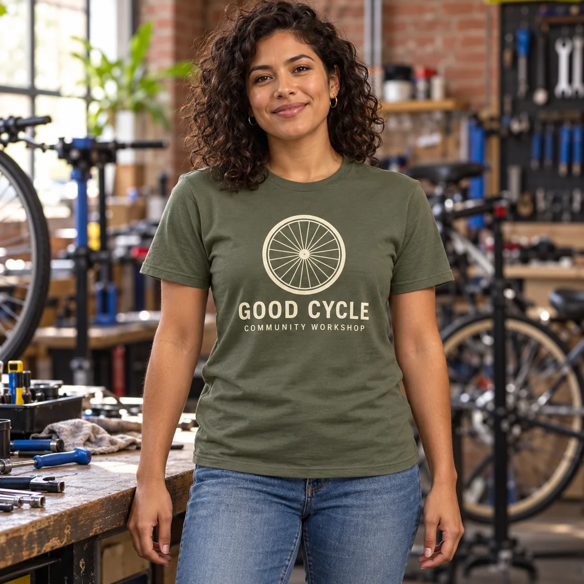 Model wearing a military green Gildan Softstyle T-shirt with sample Good Cycle artwork in a bicycle workshop.