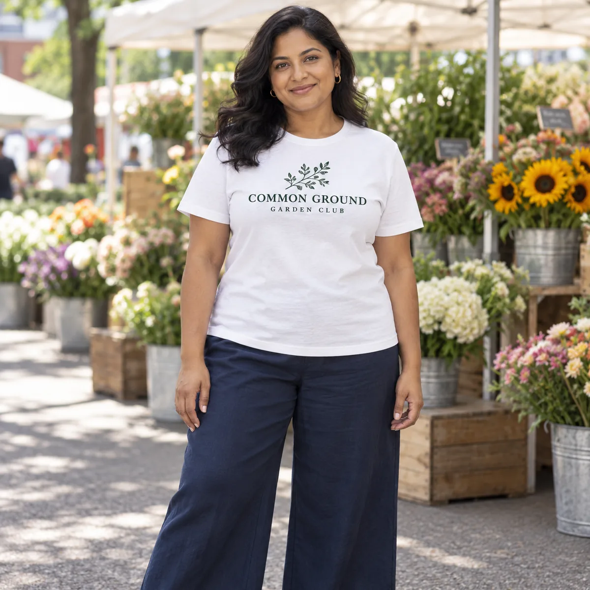 Model wearing a white Gildan Softstyle T-shirt with sample Common Ground artwork at a flower market.
