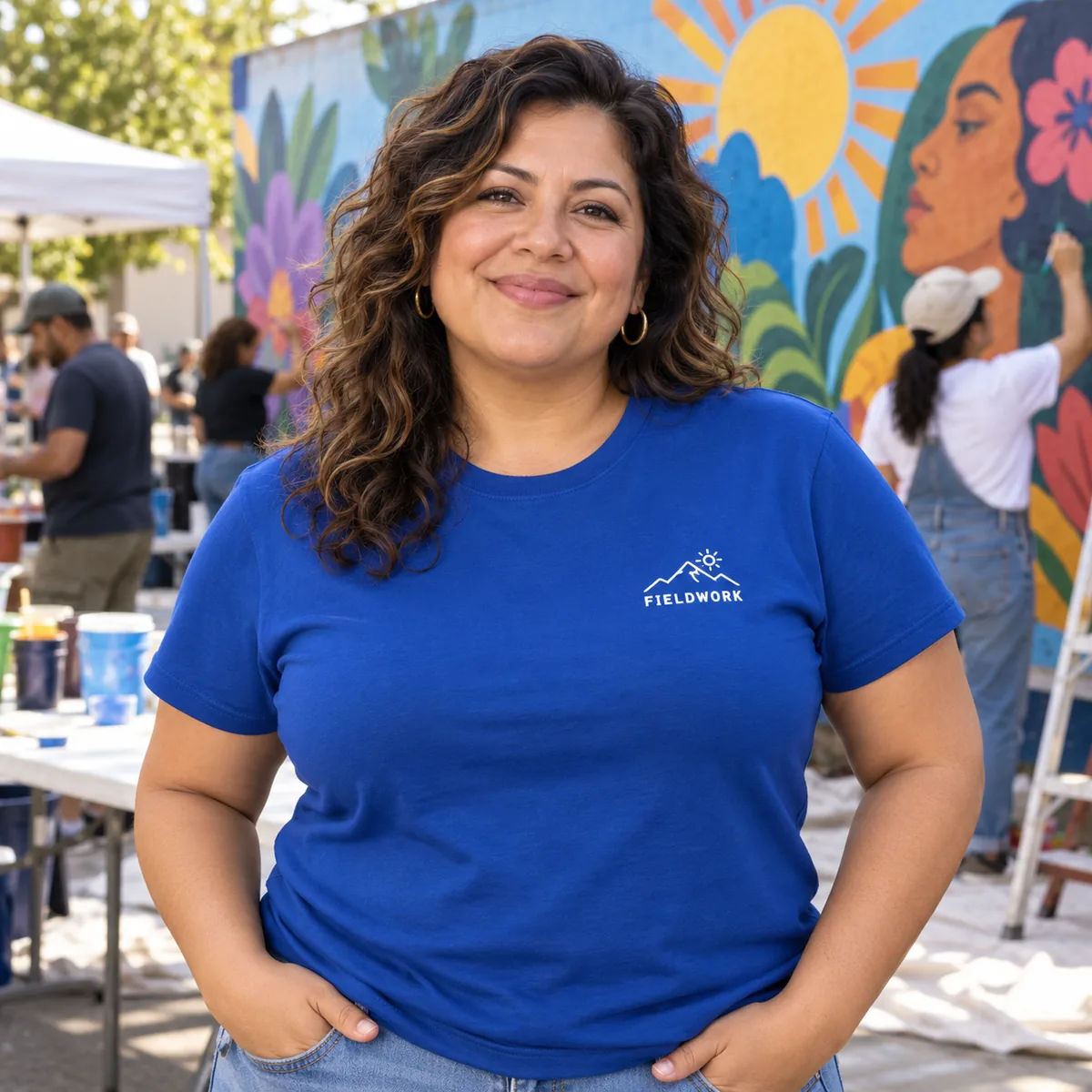 Latina woman wearing a deep royal Hanes Essential tee with fictional Fieldwork chest print at a mural event.