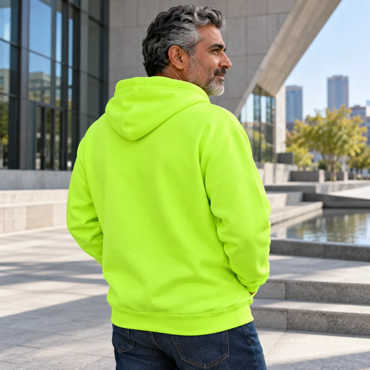Mature South Asian man wearing a safety green JERZEES NuBlend hoodie at an architecture plaza, in a back three-quarter pose.