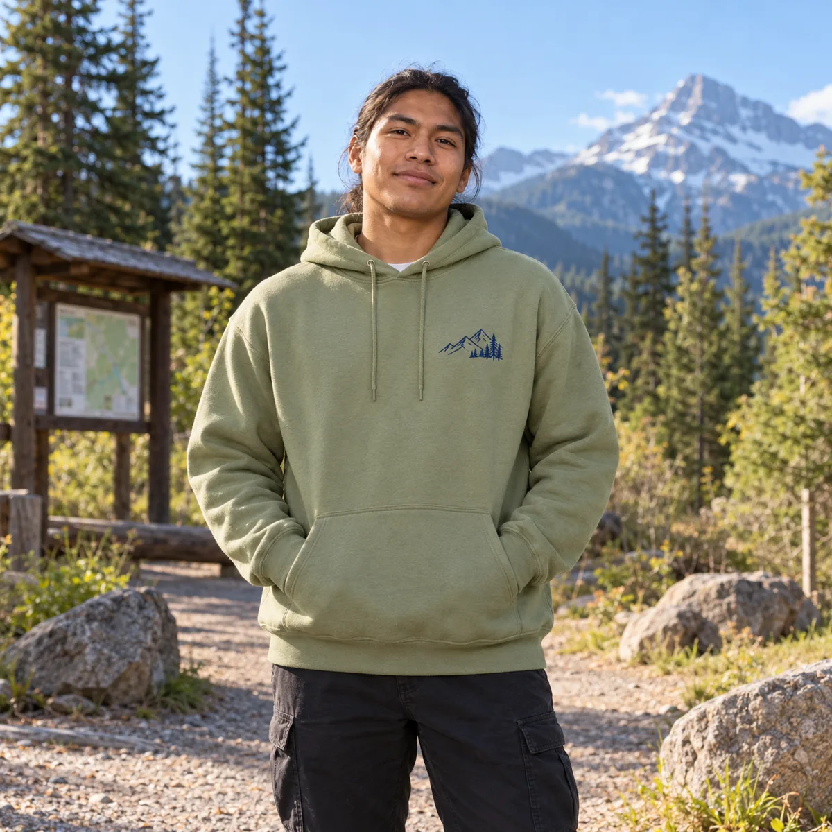 Indigenous man wearing a kiwi green hoodie with fictional navy mountain print at a forest trailhead.