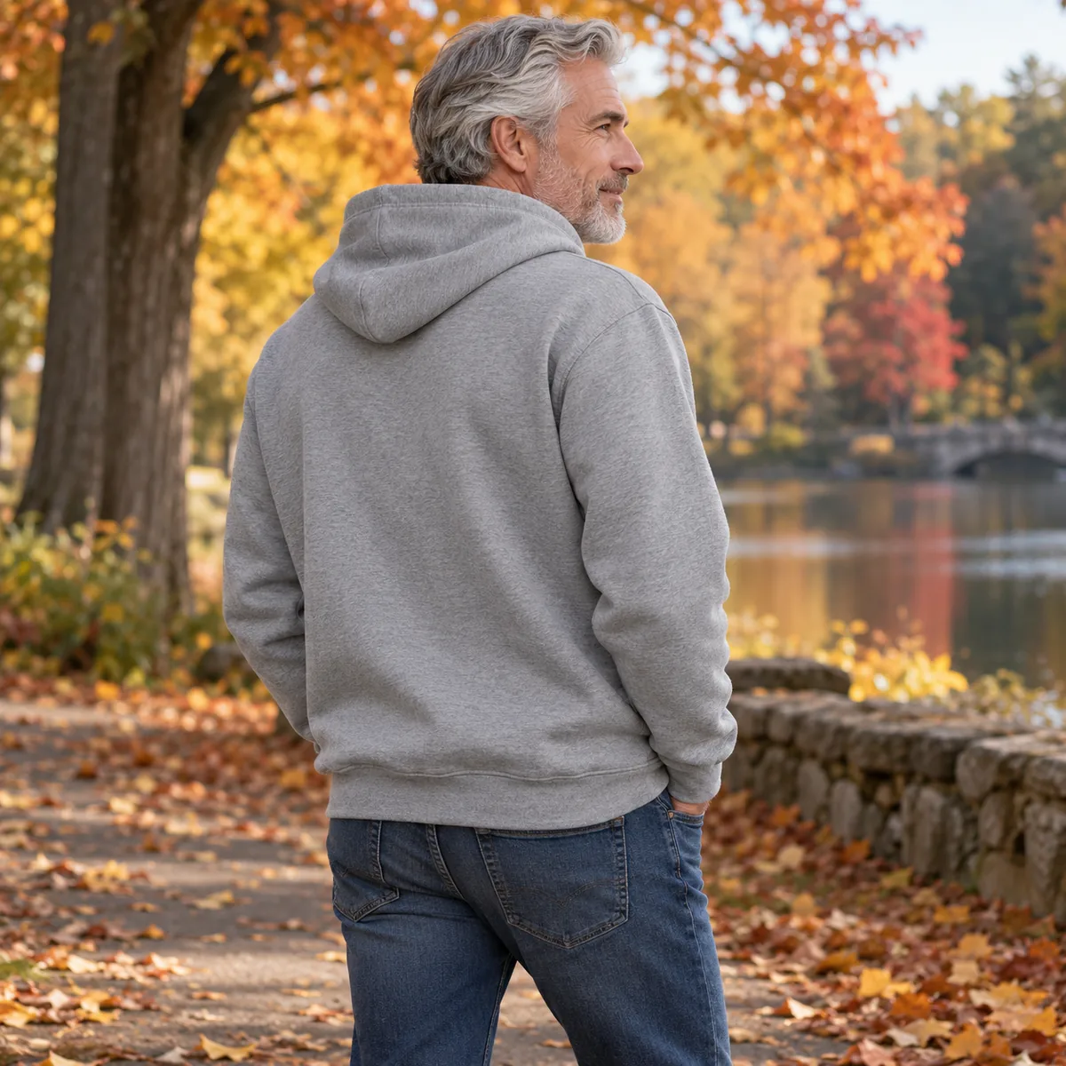 Silver-haired man wearing an Oxford grey JERZEES NuBlend hoodie in an autumn park, in back three-quarter view.