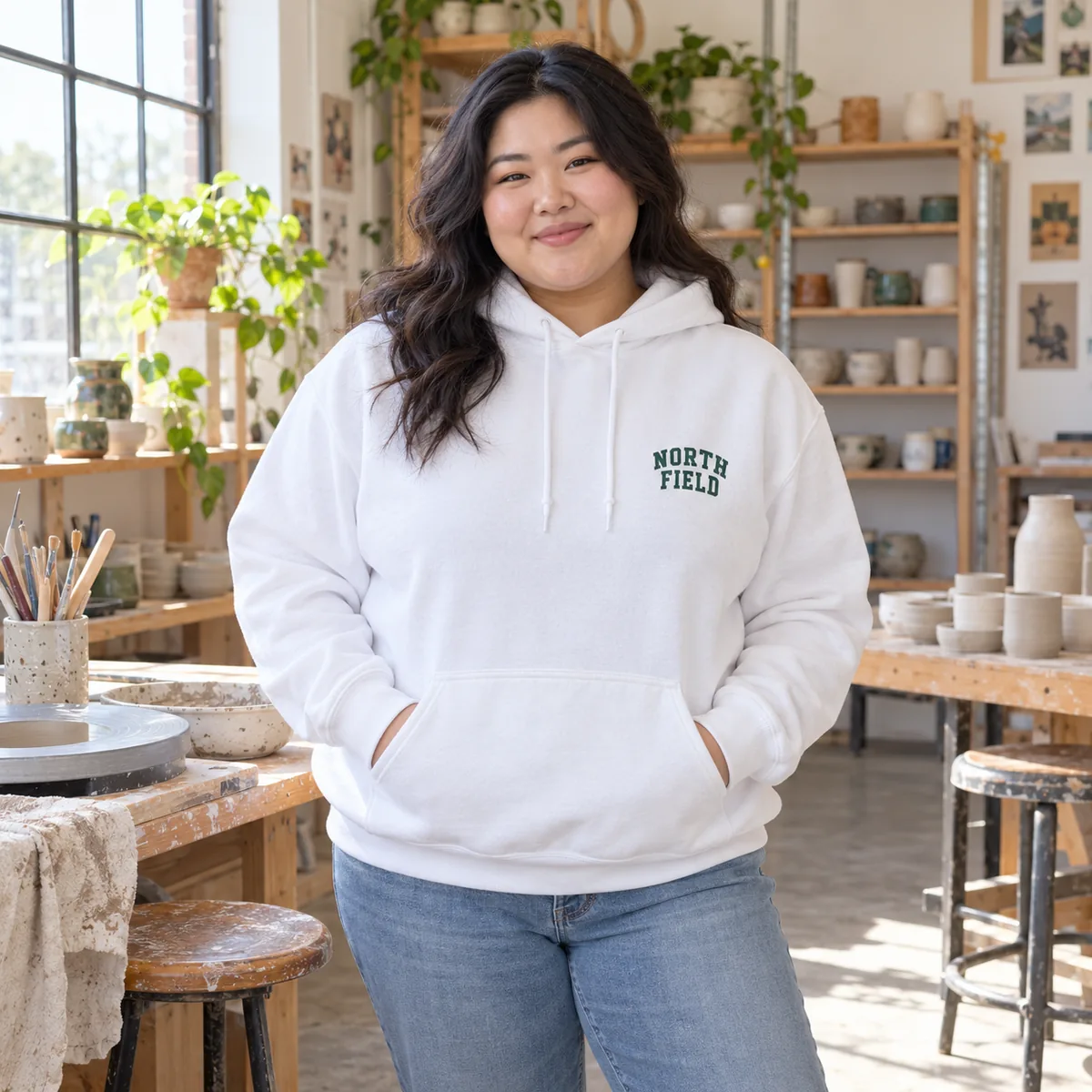 East Asian woman wearing a white JERZEES NuBlend hoodie with fictional North Field chest print at a ceramics studio.