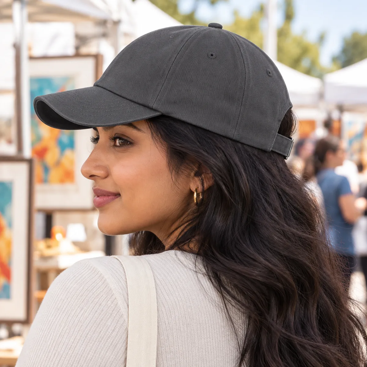 South Asian woman wearing a dark grey blank baseball cap in profile at an outdoor art fair.