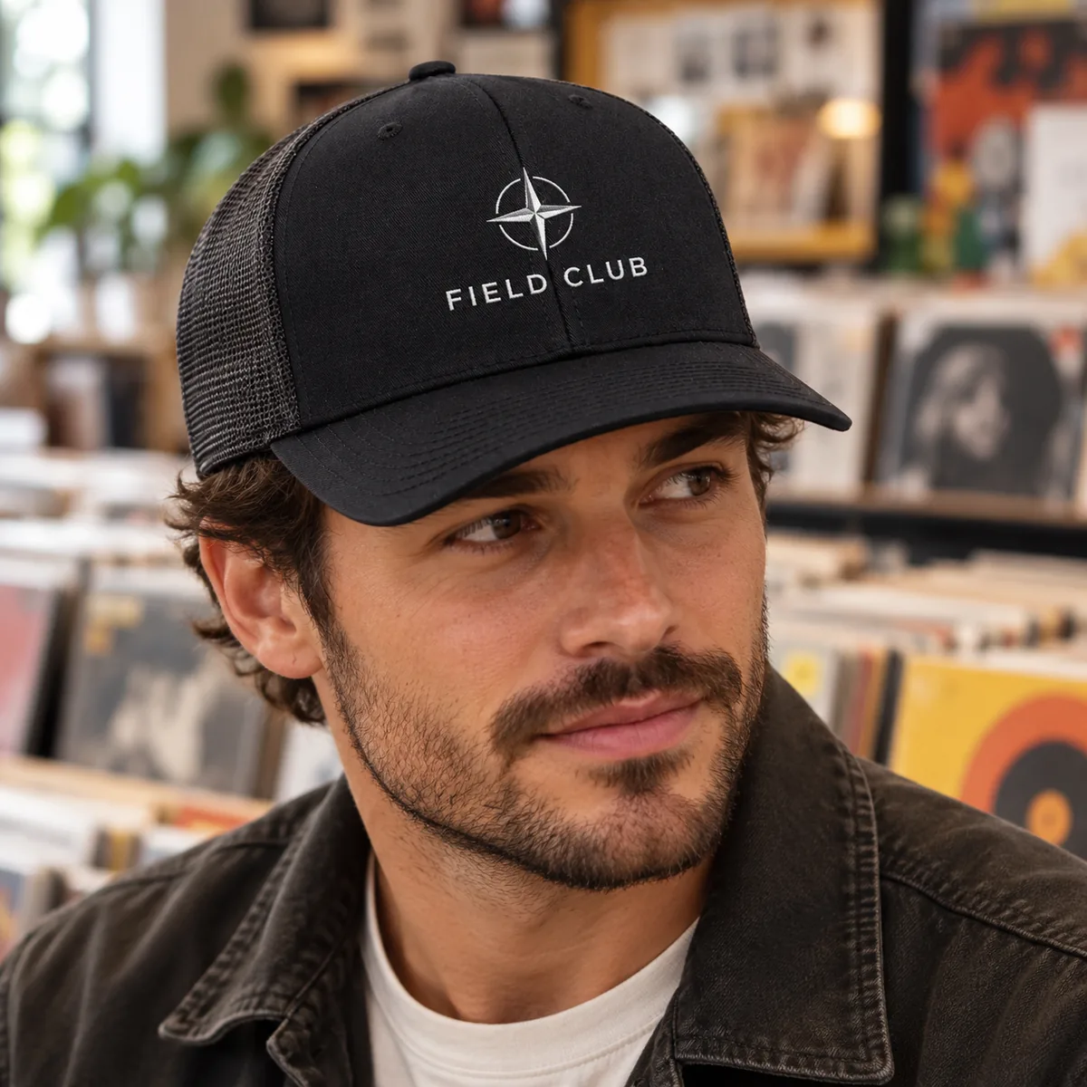 Close view of a man wearing a black Yupoong trucker cap with fictional Field Club sample embroidery at a record shop.