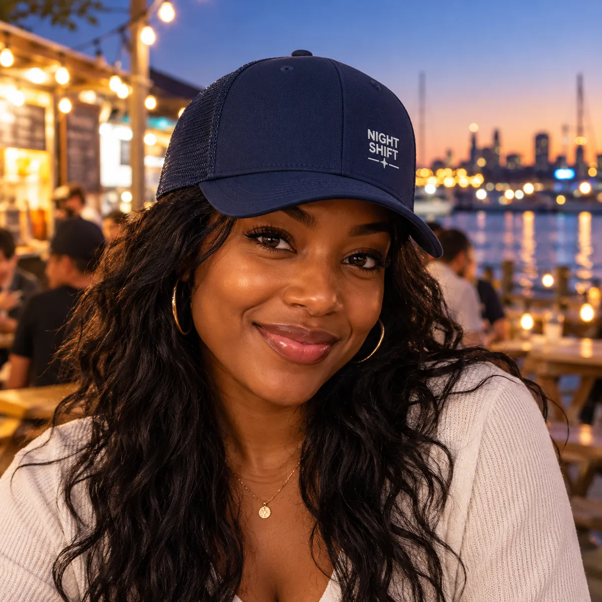 Woman wearing a navy trucker cap with fictional Night Shift sample embroidery at an evening market.