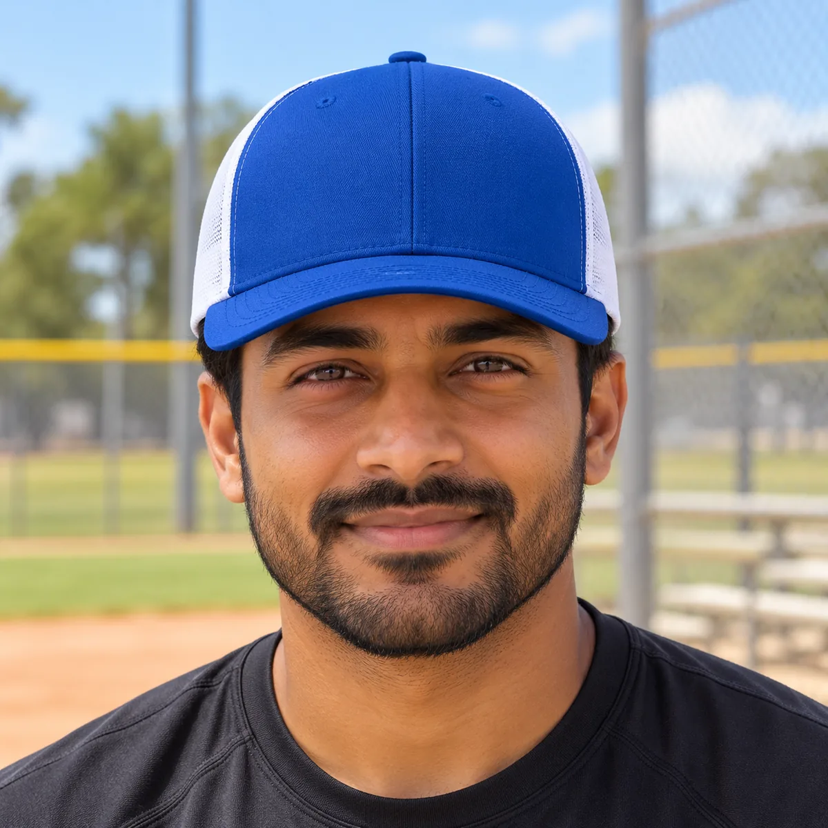 Man wearing a royal blue and white Yupoong trucker cap at a community baseball field.